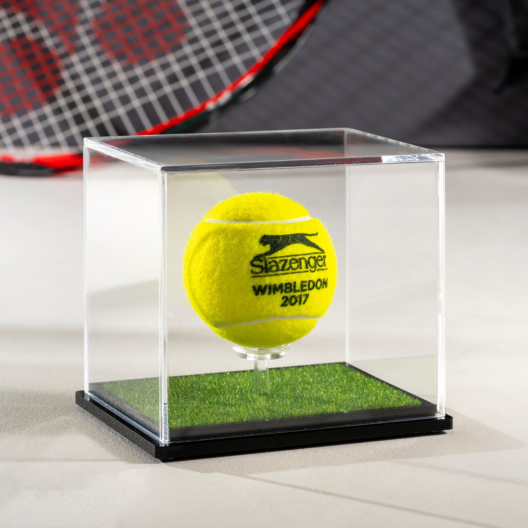 Slazenger Wimbledon 2017 tennis ball in a clear display case with white laser engraving, also a tennis racket in the background.